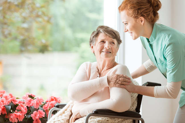 An image of a lady comforting another lady who is in a wheelchair by a window.